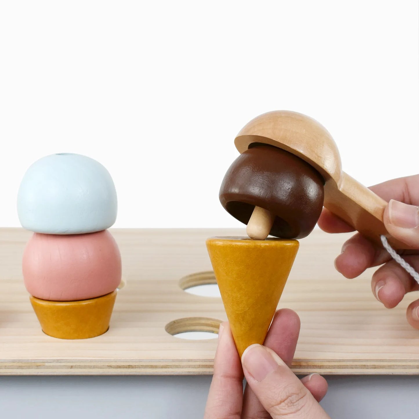 Interactive wooden ice cream toy set. Hands using a scooper to stack pretend play food scoops onto cones, perfect for toddler fine motor skills.