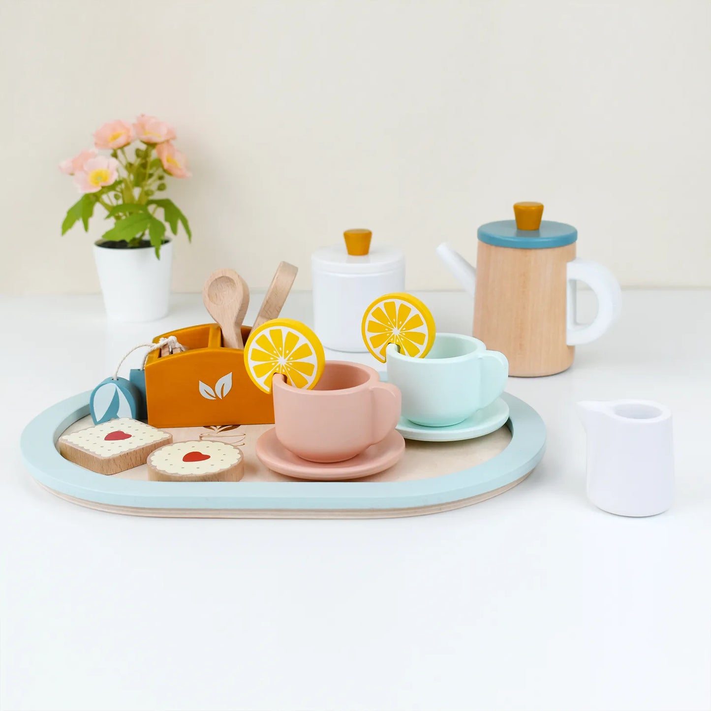 Beautifully painted wooden toy tea set for kids on a white table. This toddler pretend play set features pastel teacups, a teapot, and sweets.