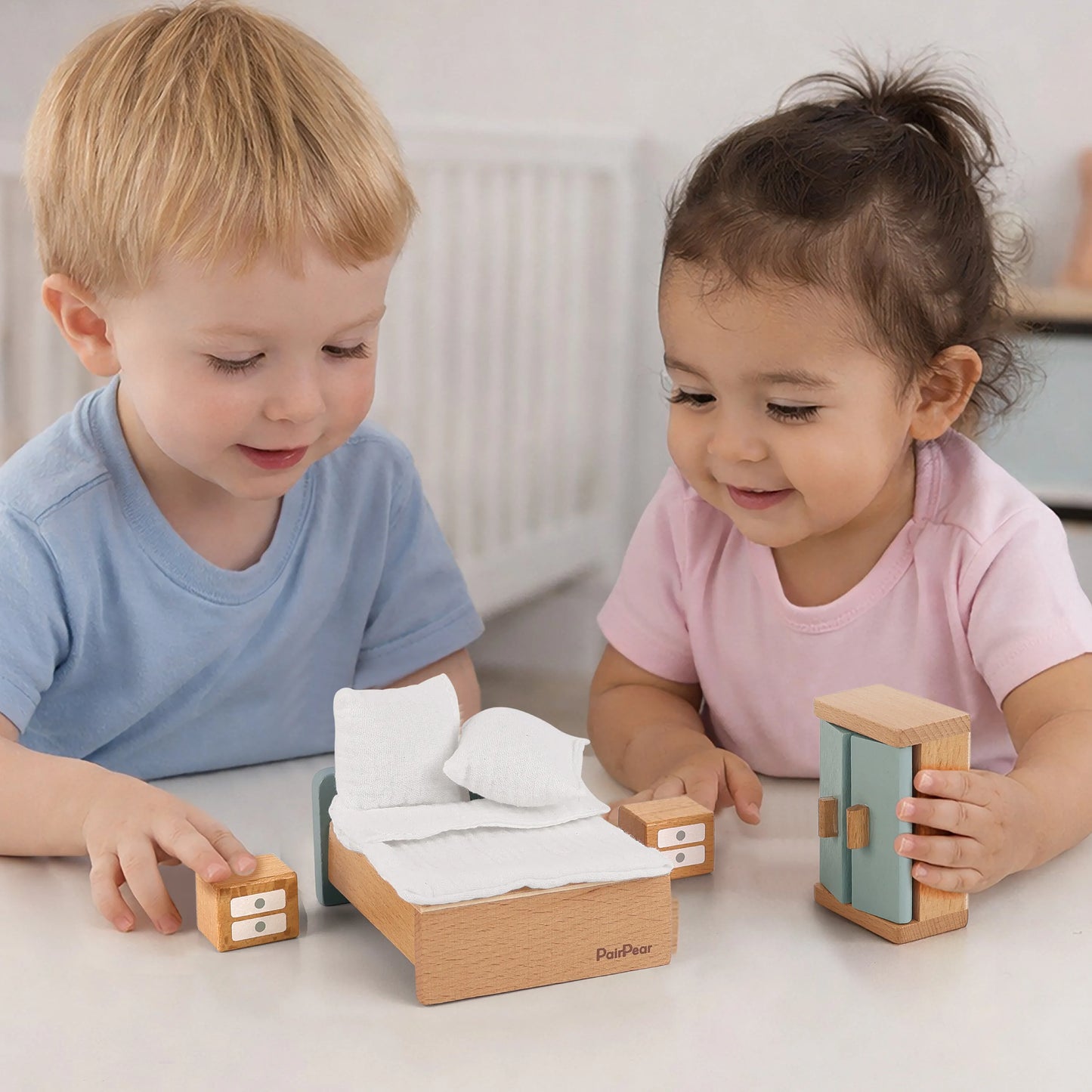 Toddler boy and girl playing with a miniature wooden bedroom furniture set. Includes a wooden bed with pillows, nightstands, and a tiny wardrobe.