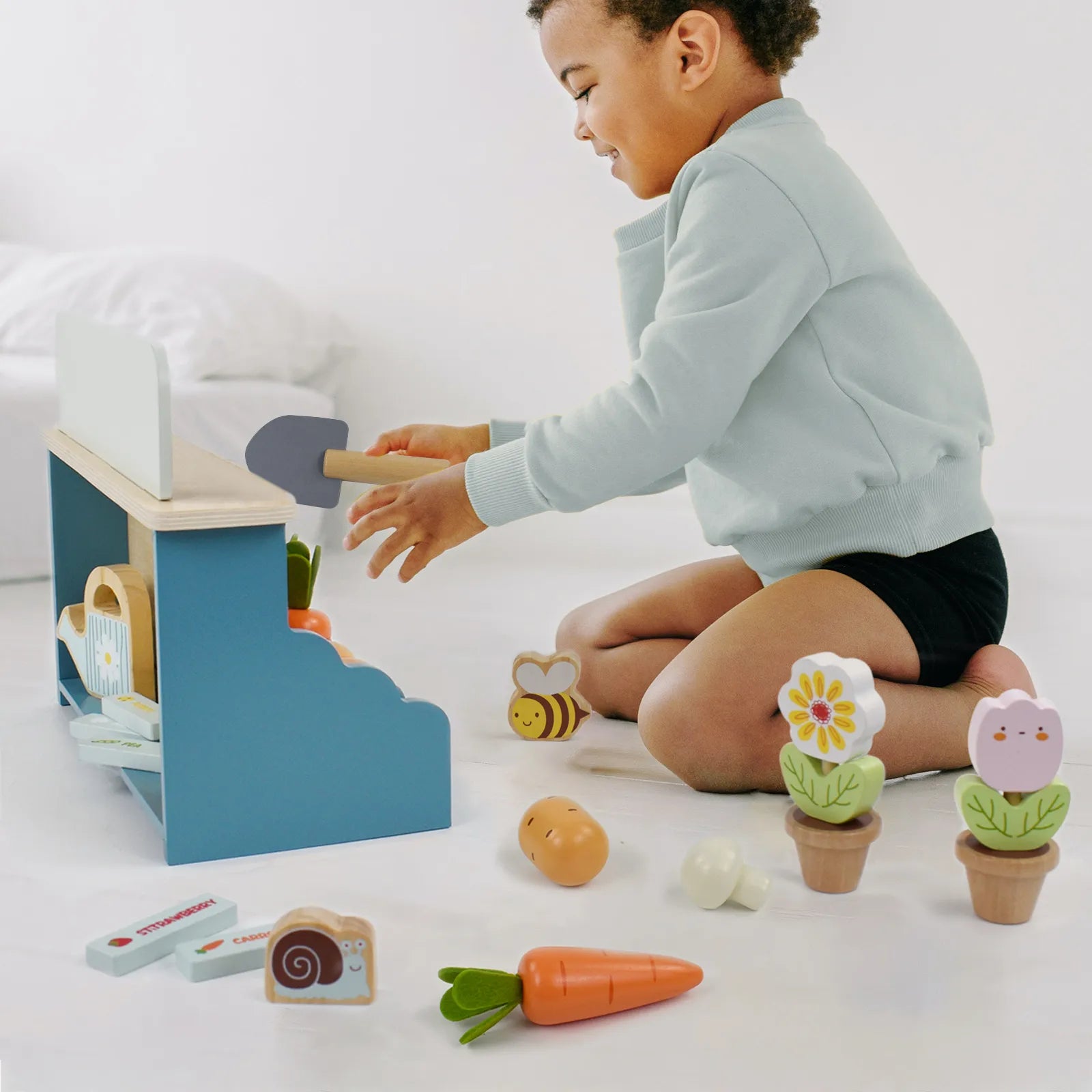 Toddler boy enjoying a pretend play garden set. He is using a toy shovel to plant wooden play food vegetables in the little farmer activity center.
