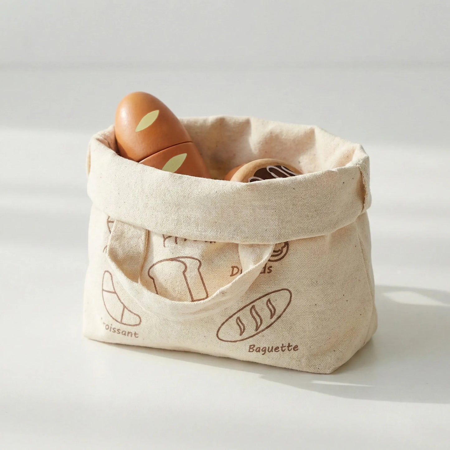 Reusable canvas bag folded into a storage basket holding toddler wooden play food. Features pretend bread and a donut from the toy bakery set.