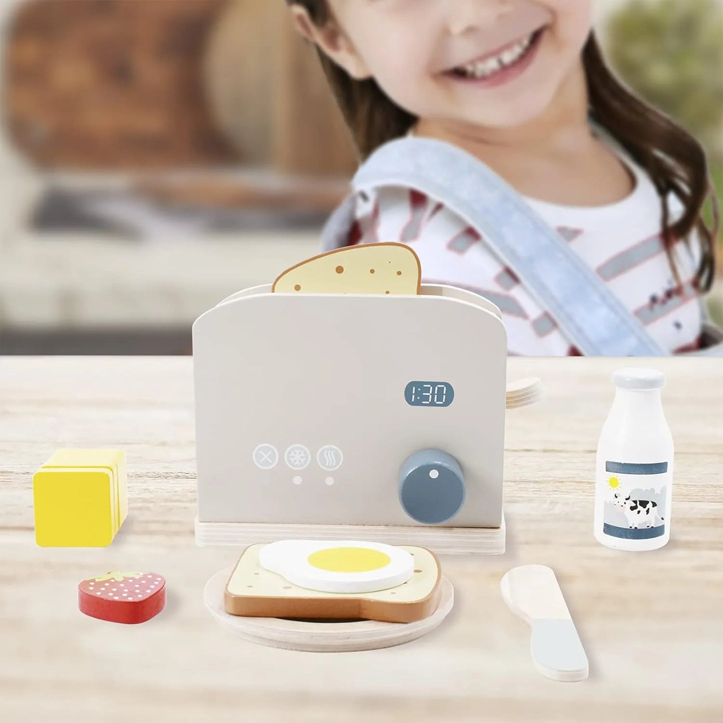 Smiling toddler playing with a wooden toaster toy set. This pretend play kitchen breakfast kit includes play food bread, egg, and milk on a table